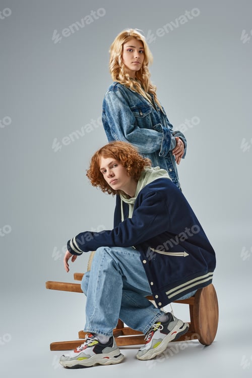 Preview: youthful couple in fashionable casual clothes looking at camera while posing near stool on grey