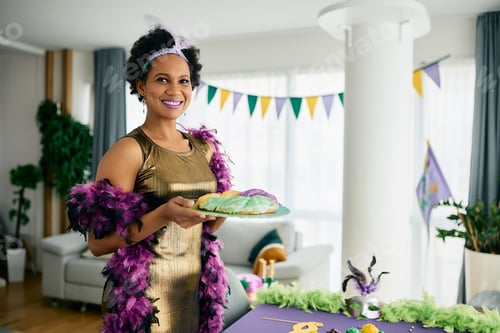 Preview: Happy African American woman in Mardi Gras costume serving King cake at home party.