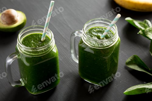 Preview: Green smoothie with avocado, spinach and banana in glass jars over black background, side view.