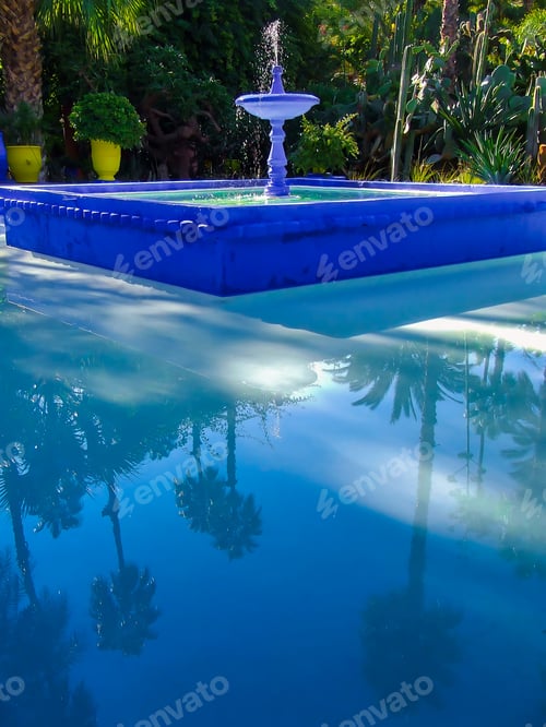 Preview: Trickling blue fountain in tropical, exotic garden with palm trees reflecting in small garden lake