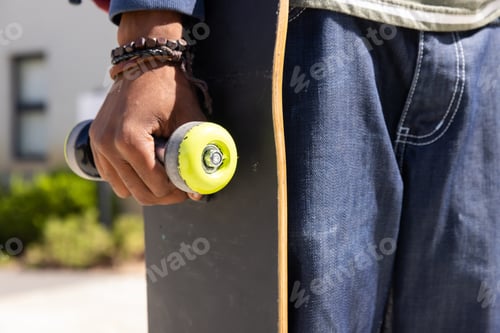 Preview: Holding skateboard, african american teenage boy wearing jeans and bracelets, hanging out outdoors