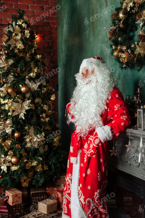 Preview: Santa Claus with bag of gifts by Christmas fireplace in decorated living room with Christmas tree