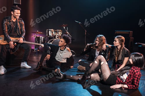 Preview: Young rock and roll band sitting together and drinking beer after concert on stage