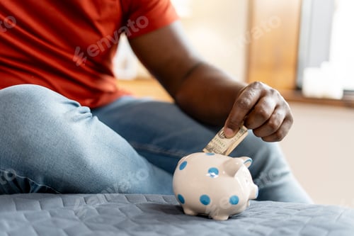 Preview: African American guy holding and putting Euro currency money into piggy bank