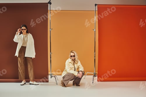 Preview: interracial women in sunglasses and autumn outerwear posing on colorful backdrop, fall fashion