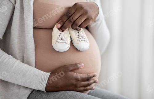 Preview: Desired Pregnancy. Black Pregnant Woman Embracing Belly And Holding Small Baby Shoes