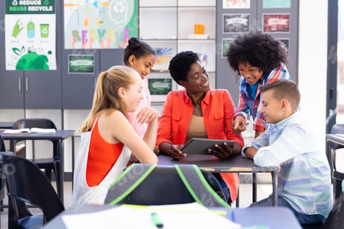 Preview: Smiling, diverse female teacher and pupils looking at tablet together in classroom