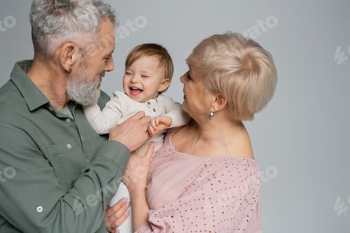 Preview: happy grandparents embracing laughing baby girl isolated on grey