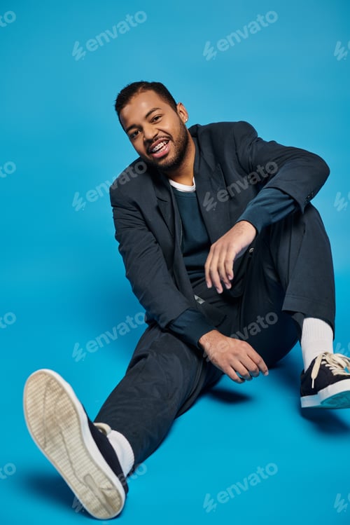 Preview: cheerful african american man sitting with bent leg and looking to camera on blue background