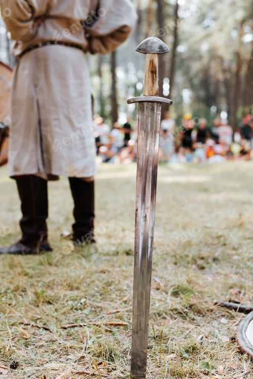 Preview: Medieval old steel sword stuck in the ground. Close-up, selective focus on the blade of the weapon