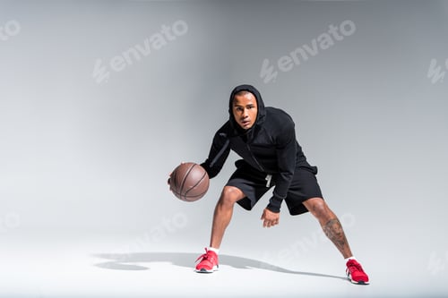 Preview: sporty african american man playing with basketball ball and looking at camera on grey