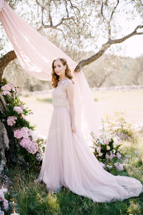 Preview: Bride in a long dress stands near a tree decorated with flowers