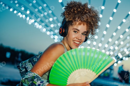 Preview: Young woman listening music with headphones and cooling with hand fan at summer festival
