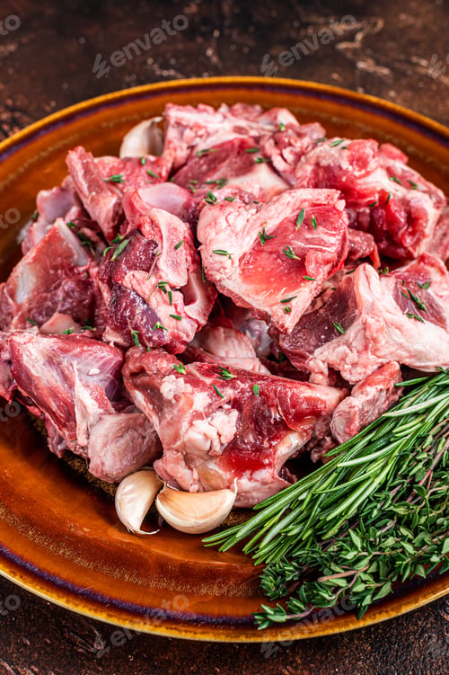Preview: Raw mutton meat diced for goulash or stew with bone on a rustic plate. Dark background. Top view
