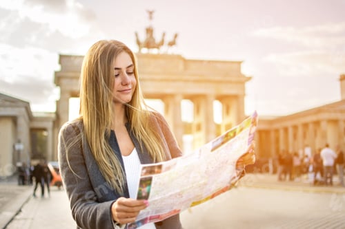 Preview: Beautiful young woman looking at map guide while standing in front of Brandenburg gate at sunset.