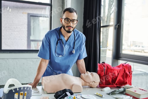 Preview: bearded paramedic in eyeglasses and blue uniform looking at camera near CPR manikin, red first aid