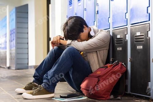 Preview: Sitting by school lockers, asian teenage boy resting head on arms with backpack and notebook