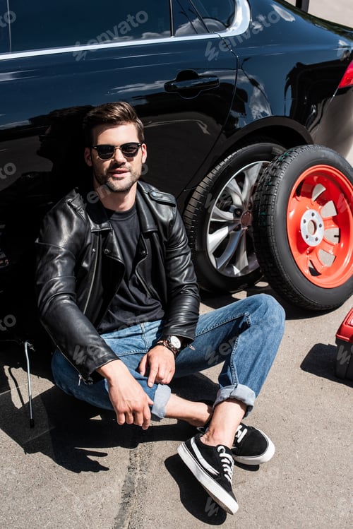 Preview: smiling stylish man in sunglasses sitting near broken car at street