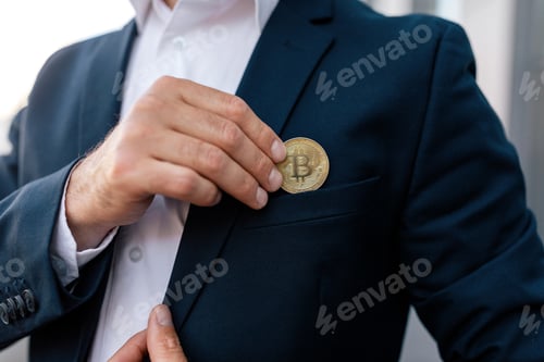 Preview: Confident young caucasian guy ceo manager in suit puts bitcoin coin in pocket near modern outdoor