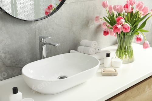 Preview: Vase with beautiful pink tulips and toiletries near sink in bathroom