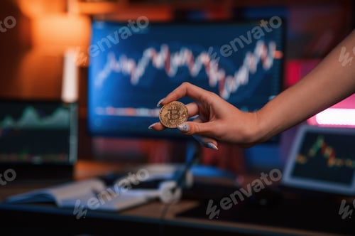 Preview: Close up view of woman's hand that holding bitcoin against computer display and tablet with graphs