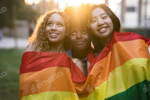 Preview: Three gay multiracial female friends are standing outdoors with a pride flag on a sunny summer day