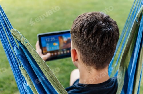 Preview: Rear view of gen-z boy sitting in hammock chair in back yard, using tablet computer