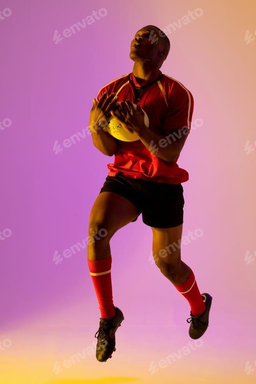 Preview: African american male rugby player with rugby ball over pink lighting