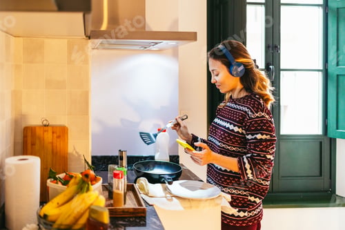 Preview: Woman cooking and having breakfast while listening music with headphones in kitchen