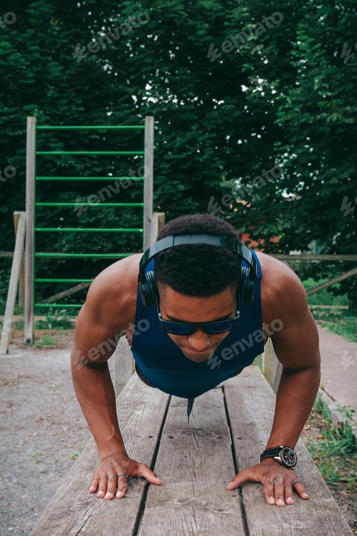 Preview: Young fit man with sunglasses and headphones doing push-ups in the park