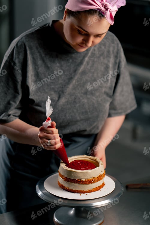 Preview: female baker in a professional kitchen puts red cream from a pastry bag onto a sponge cake
