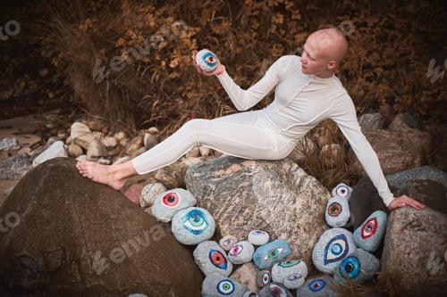 Preview: Young hairless girl with alopecia in white futuristic costume pensively examines stone with eye