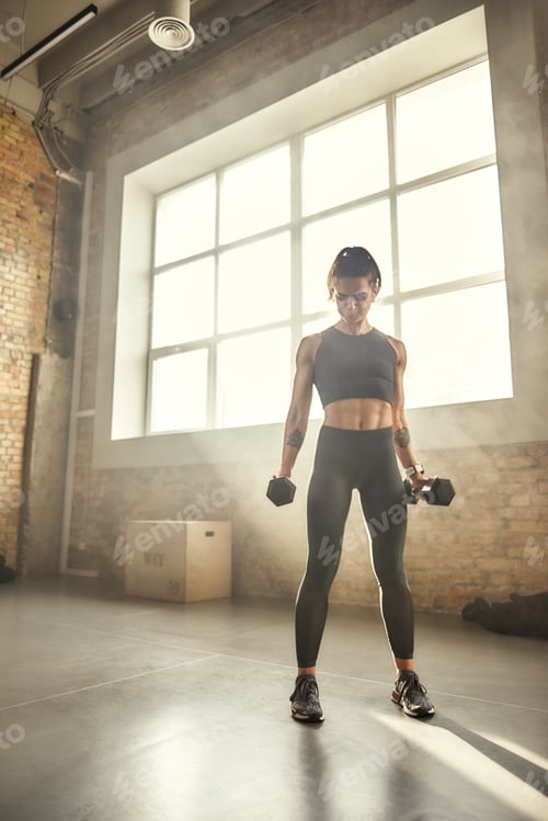 Preview: Working with weight. Young athletic woman in sportswear exercising with dumbbells while standing in