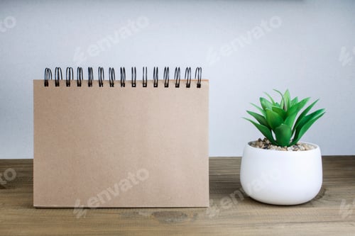 Preview: An open notebook with blank pages next to a green plant in a pot on a wooden table.
