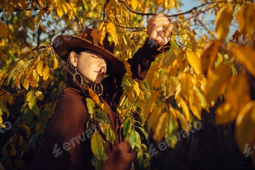 Preview: Stylish woman in hat and brown clothes posing among autumn leaves in warm sunny light