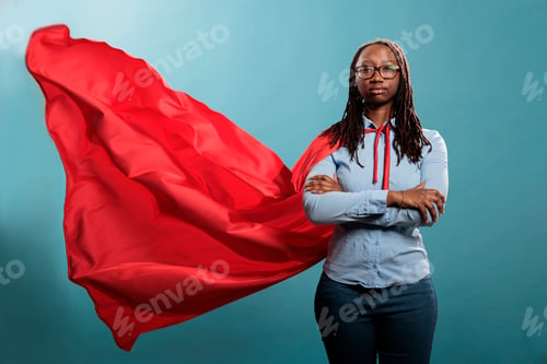 Preview: Confident and brave young superhero woman standing with arms crossed while wearing red mighty cape
