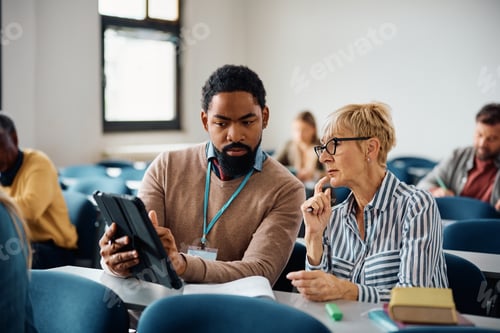 Preview: Black teacher using touchpad while assisting senior woman with lecture in the classroom.
