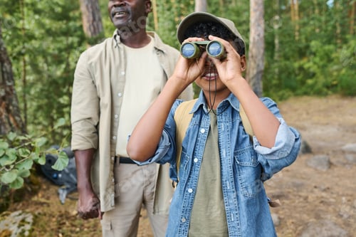 Preview: Cute little boy in casualwear and baseball cap looking through binoculars