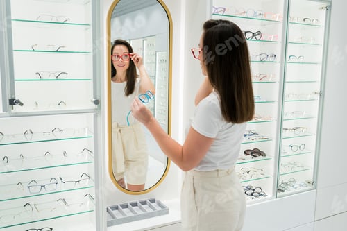 Preview: Young smiling woman choosing eyeglasses at optical store.
