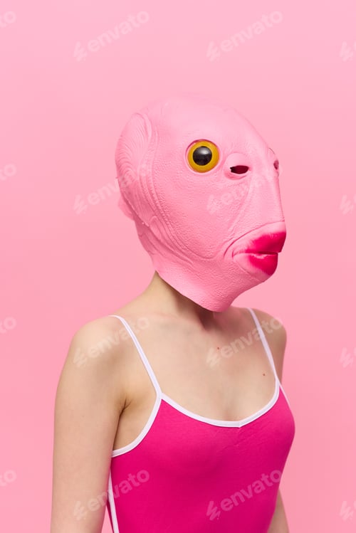 Preview: Funny crazy woman on a pink background standing in a fish head mask on a pink background, conceptual