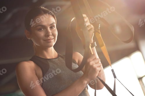 Preview: Portrait of a young attractive woman adjusting trx fitness straps, looking at camera and smiling