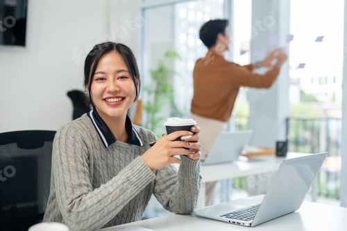 Preview: A charming Asian businesswoman sits at her desk in the office with a coffee cup in her hand.