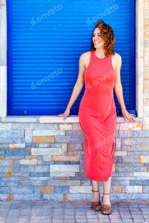 Preview: Woman in sleeveless red dress standing near bright wall