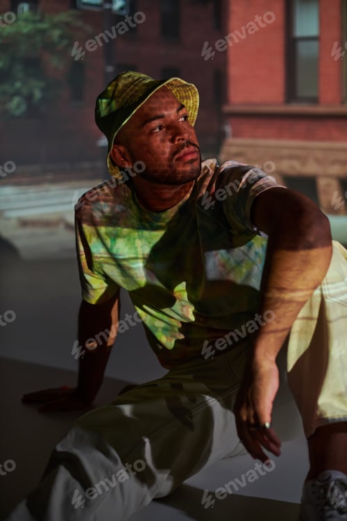 Preview: handsome male model sitting on floor in vibrant outfit in digital projector lights, fashion concept