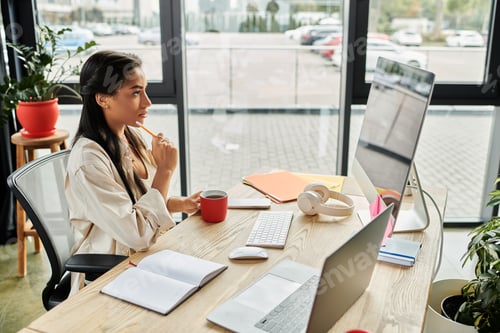 Preview: Young woman engaged in creative thinking at her modern office desk with plants and technology