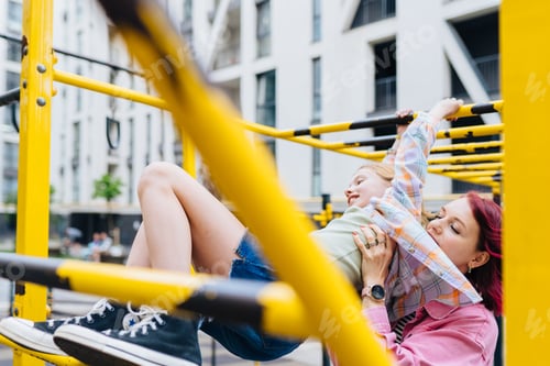 Preview: Pink haired mother helping teen daughter across monkey bars, low angle view.
