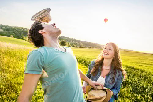 Visualização: Casal despreocupado brincando com um chapéu em um campo ensolarado com um balão de ar quente à distância