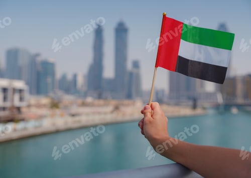 Preview: female hand holding tiny flag of UAE against Dubai skyline.