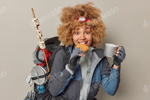 Preview: Indoor shot of pretty woman hiker has snack during journey bites waffle and drinks tea dressed in co