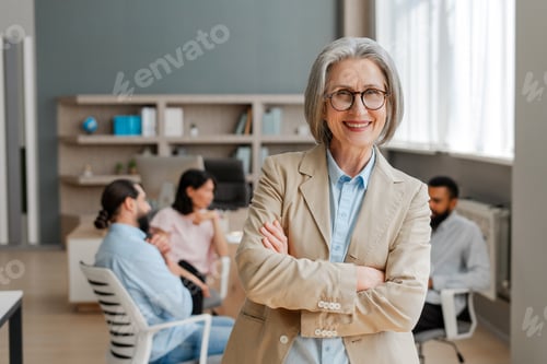 Preview: Senior businesswoman smiling with crossed arms in busy office looking at camera